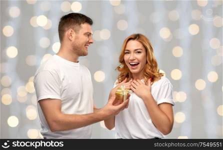 relationships, holiday and valentines day concept - happy couple in white t-shirts with christmas gift over festive lights background. happy couple in white t-shirts with christmas gift