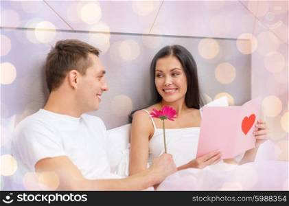 relationships, holidays, valentines day and love concept - smiling couple in bed with postcard and pink flower over lights background