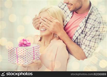relationships, love, people, birthday and holidays concept - happy man covering woman eyes and giving gift box over lights background