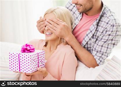 relationships, love, people, birthday and holidays concept - happy man covering womans eyes and giving gift box at home