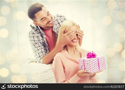 relationships, love, people, birthday and valentines day concept - happy man covering woman eyes and giving gift box at home over holidays lights background