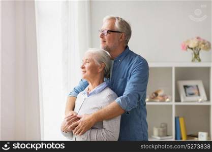 relationships, old age and people concept - happy senior couple looking through window and hugging at home. happy senior couple looking through window at home