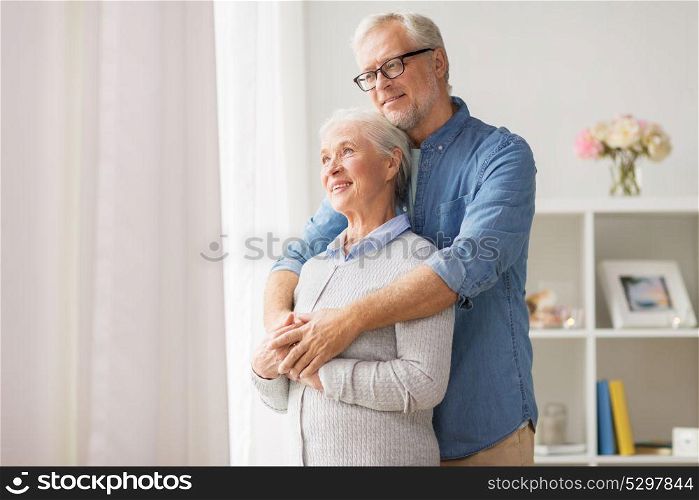 relationships, old age and people concept - happy senior couple looking through window and hugging at home. happy senior couple looking through window at home