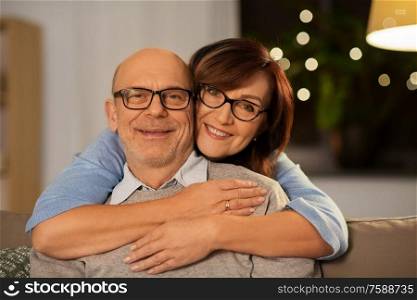 relationships, old age and people concept - happy smiling senior couple hugging on sofa at home in evening. happy senior couple hugging on sofa at home