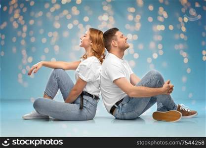 relationships, style and people concept - happy couple in white t-shirts sitting on floor over lights on blue background. happy couple in white t-shirts sitting on floor