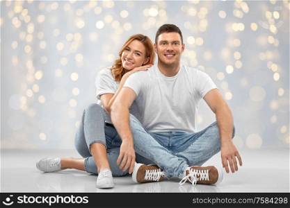 relationships, style and people concept - happy couple in white t-shirts sitting on floor over festive lights background. happy couple in white t-shirts sitting on floor