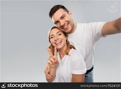 relationships, technology and people concept - happy couple in white t-shirts taking selfie smartphone and showing peace by over grey background. happy couple in white t-shirts taking selfie