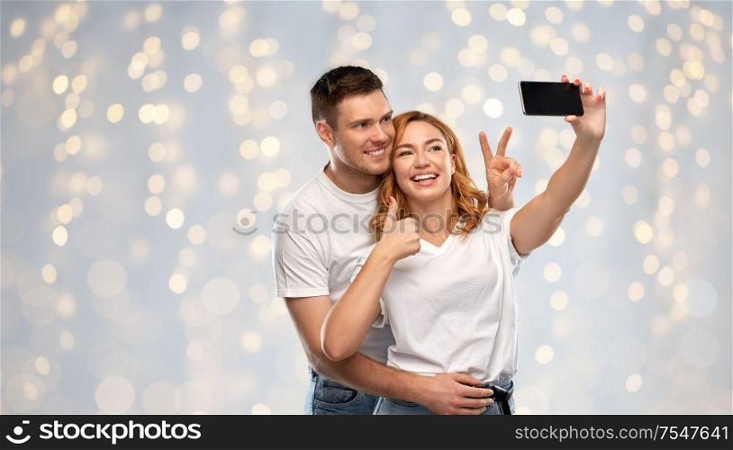 relationships, technology and people concept - happy couple in white t-shirts taking selfie smartphone and showing peace by over holidays lights background. happy couple in white t-shirts taking selfie