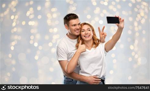 relationships, technology and people concept - happy couple in white t-shirts taking selfie smartphone and showing peace by over holidays lights background. happy couple in white t-shirts taking selfie