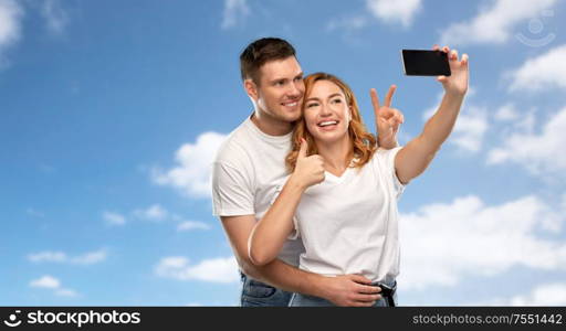 relationships, technology and people concept - happy couple in white t-shirts taking selfie smartphone and showing peace by over blue sky and clouds background. happy couple in white t-shirts taking selfie