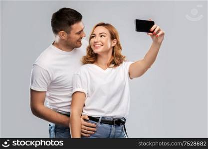 relationships, technology and people concept - portrait of happy couple in white t-shirts taking selfie smartphone by over grey background. happy couple in white t-shirts taking selfie