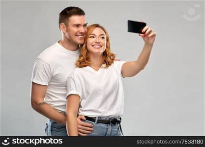relationships, technology and people concept - portrait of happy couple in white t-shirts taking selfie smartphone by over grey background. happy couple in white t-shirts taking selfie
