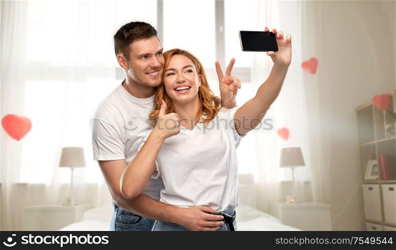 relationships, technology and valentine&rsquo;s day concept - happy couple in white t-shirts taking selfie smartphone and showing peace by over bedroom decorated with heart shaped balloons background. happy couple taking selfie on valentines day