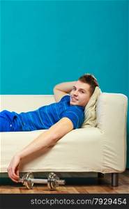 Relax after sport activity. Young man fit body relaxing on couch, dumb bell on floor