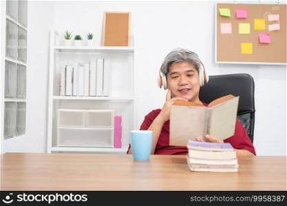 Relax elderly Asian man reading a book and listening to music in home office.