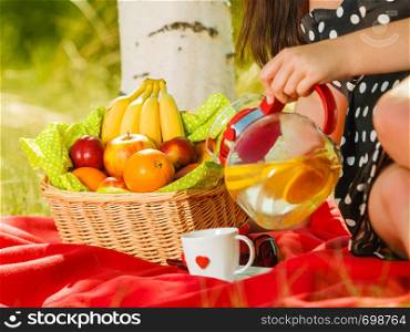 Relaxation during summertime concept. Picnic basket with fruit on red blanket in woods, woman pouring orange water into cup.. Picnic basket on blanket, woman pouring water into cup
