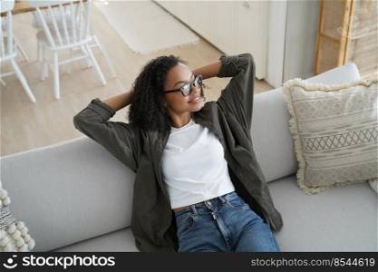 Relaxed african american girl sitting on comfortable sofa in living room, hands behind head. Biracial young woman relaxing on couch at home, dream, enjoying weekend. Stress free, wellness.. Relaxed african american girl rest, sit on comfortable couch in living room. Stress free, wellness