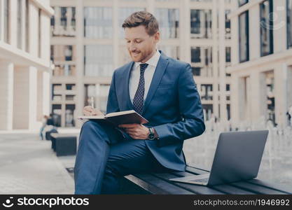 Relaxed and confident office worker in formal blue suit working outside while sitting on bench, using laptop and writing notes in agenda, city buildings in blurred background. Relaxed confident office worker in formal blue suit writing notes in agenda while working outside