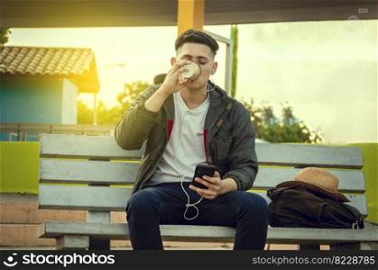 Relaxed attractive boy listening to music on a bench, boy sitting on a bench listening to music and drinking coffee
