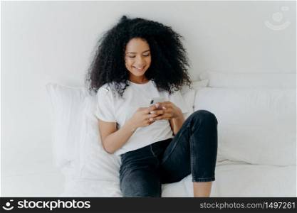 Relaxed beautiful Afro woman with cheerful expression uses modern mobile phone, surfs internet, poses in white comfortable bed, connected to wifi, browses webpage, enjoys domestic atmosphere