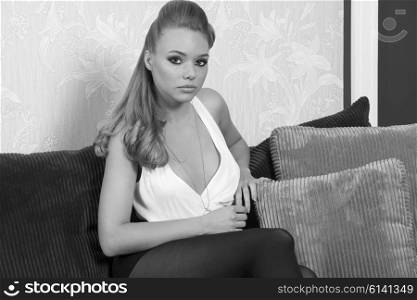 relaxed beautiful blonde girl with cute make-up, long blonde hair and elegant fashion clothes sitting on sofa and looking in camera , in black and white