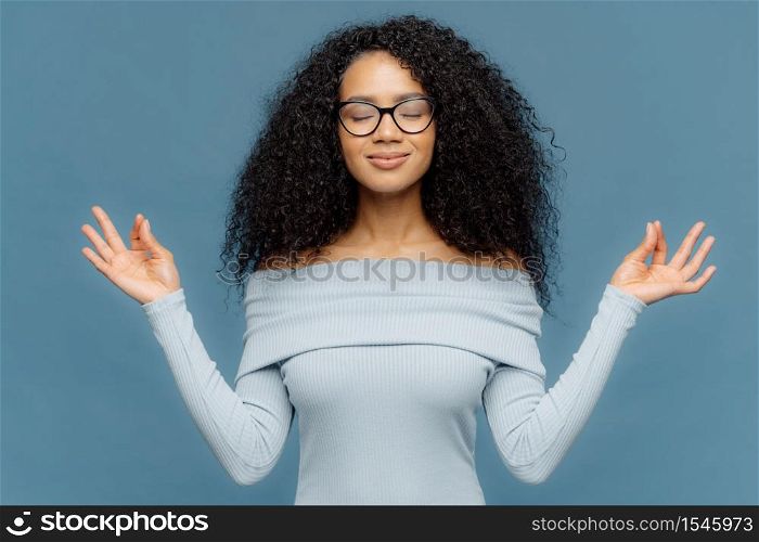 Relaxed beautiful woman with Afro hairstyle, feels relieved, shows zen gesture, keeps hands in okay gesture, controls emotions, gets in control with herself has eyes closed isolated on blue background