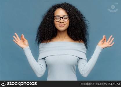 Relaxed beautiful woman with Afro hairstyle, feels relieved, shows zen gesture, keeps hands in okay gesture, controls emotions, gets in control with herself has eyes closed isolated on blue background