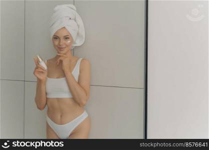 Relaxed beautiful young woman applying face cream at home, using cosmetic product for healthy skin while posing with smile in bathroom, wearing wrapped towel on head. Beauty treatment concept. Relaxed beautiful young woman applying face cream in bathroom, using cosmetic product for healthy skin