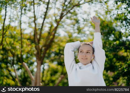 Relaxed beautiful young woman does stretching exercises outdoors keeps arms raised up focused into distance wears white hoodie listens music in earphones has training in park during sunny day