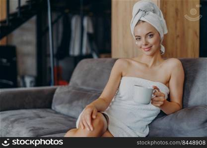 Relaxed beautiful young woman has healthy skin wears soft towel on head and around naked body holds mug of coffee poses at sofa with aromatic beverage enjoys weekends has well groomed complexion