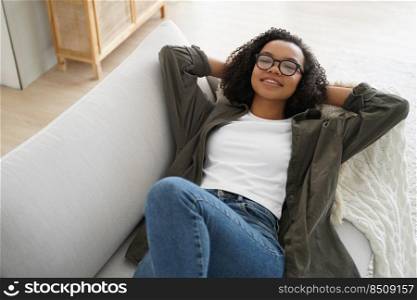 Relaxed biracial teen girl in glasses lying on comfortable couch, hands behind her head, relax on sunny lazy weekend at home. Serene young woman relaxing on cozy sofa in living room enjoy break.. Relaxed biracial teen girl in glasses rest, lying on couch, enjoy break on sunny weekend at home