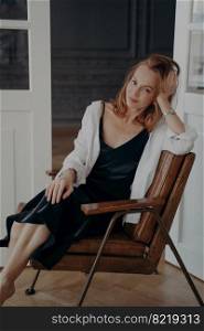 Relaxed blond woman in black evening dress and white shirt. Stylish confident mid adult lady is sitting in an arm chair. Happy european woman is sitting have her head leaned to the hand after party.. Relaxed blond woman in black evening dress. Stylish confident mid adult lady is sitting in chair.