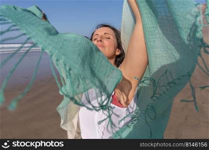 Relaxed brunette raising arms with green scarf and enjoying sunlight with closed eyes while spending time on sandy beach during summer vacation. Female traveler with scarf enjoying sunlight on beach