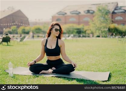 Relaxed brunette woman sits on fitness mat on lotus yoga pose wears sunglasses cropped top and leggings looks aside drinks water after training breathes deeply fresh air. Healthy lifestyle concept