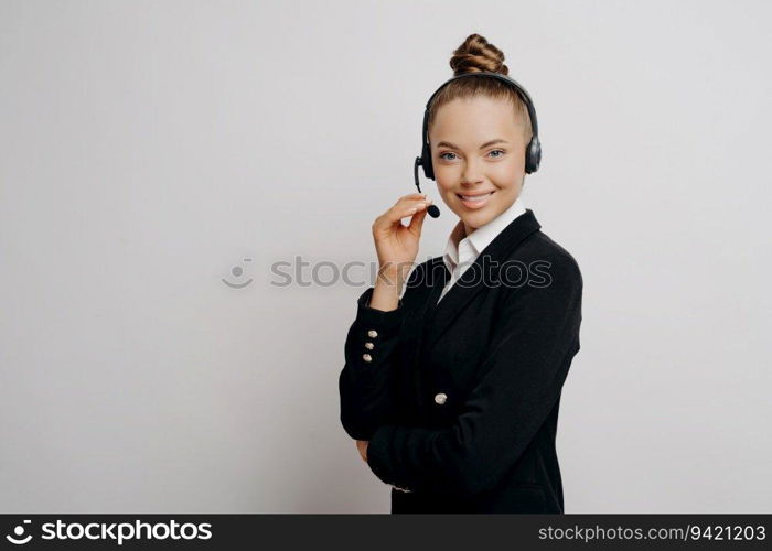 Relaxed businesswoman in dark suit, wireless headset, poses positively during web conversation with colleagues in studio.