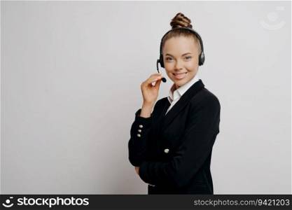 Relaxed businesswoman in dark suit, wireless headset, poses positively during web conversation with colleagues in studio.