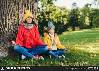 Relaxed carefree woman and little daughter sit in pose of lotus near tree in park, close eyes, try to concentrate, practice yoga together, dressed in warm clothes. Two female think about piece outdoor