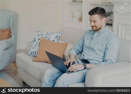 Relaxed casually dressed young businessman working from home while sitting on comfortable couch in light living room, typing on laptop and using special business software. Freelance concept. Young businessman working from home while sitting on couch