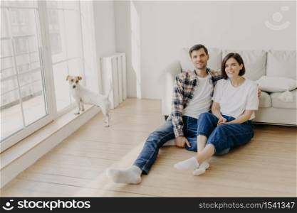 Relaxed couple sit on floor near couch, embrace and smile, dressed in casual clothes and white socks, enjoy domestic atmosphere, their dog stands near balcony window in empty spacious living room