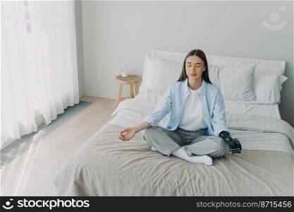 Relaxed disabled woman sit on bed in lotus position, does mudra gesture with bionic prosthetic hand, practicing yoga, relieving stress. Serene female with disability meditates with closed eyes at home. Disabled girl practicing yoga on bed, does mudra gesture with bionic prosthetic hand. Stress relief