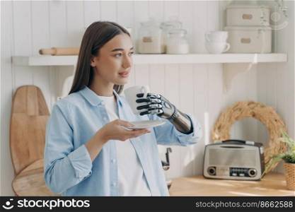 Relaxed disabled young girl holds cup of coffee, using bionic prosthetic arm, enjoying daily routine at home. Modern girl with disability holds grasped mug in artificial hand, standing in kitchen.. Relaxed disabled girl holds cup of coffee, using bionic prosthetic arm, enjoy daily routine at home