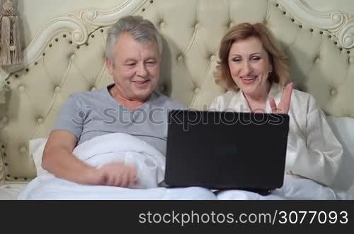 Relaxed elderly couple lying in bed in the morning and chatting with their friends through internet using laptop computer. Mature husband and wife connecting with family via skype on laptop and communicating.