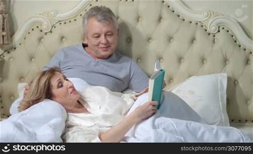 Relaxed elderly woman lying down on her back with her head on husband&acute;s lap and reading a book in bed while retired man caressing her hair. Mature couple enjoying time together in bedroom and lounging.