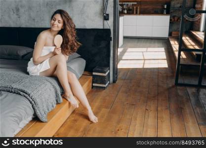 Relaxed european woman enjoys her long shiny hair and combs it with wooden brush. Brunette girl wrapped in towel after bathing has hair care routine sitting on bed. Hair cosmetics applying.. European woman enjoys her long shiny hair and combs it with wooden brush sitting on bed.