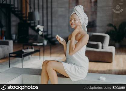 Relaxed European woman has natural beauty demonstrates slender legs applies nourishing face lotion, takes care of complexion, enjoys hygiene treatments at home, poses against cozy room interior