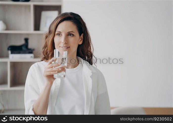 Relaxed european woman is drinking transparent pure water at home. Happy girl is holding glass and smiling. Filtered water for healthy lifestyle. Morning routine for beauty and wellness.. Relaxed european woman is drinking pure water at home. Happy girl is holding glass and smiling.