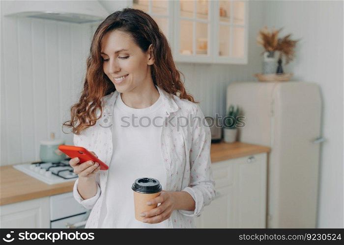 Relaxed european woman with smartphone has coffee. Girl is reading message in her kitchen at home and smiling. Modern white scandinavian interior. Stove, worktop, refrigerator and cuisine.. European woman with smartphone has coffee. Girl is reading message in kitchen at home and smiling.