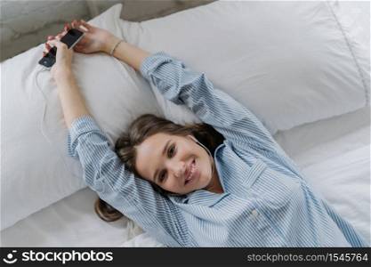 Relaxed European woman with toothy enjoys good rest in bed, stretches during morning, holds modern cell phone, listens favourite song with earphones, rejoices start new day. Horizontal view.