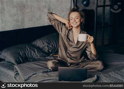 Relaxed female freelancer having cup of coffee stright after waking up in bed while checking new messages and emails on laptop, dressed in pajama. Woman with closed eyes ready to start remote work. Relaxed young brunette female in satin pajama having her cup of coffee in bed and checking emails on laptop