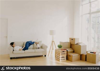 Relaxed female lies on white comfortable sofa with dog, feels tired after relocation and carrying boxes with belongings, l&stands near, dressed in casual white t shirt, jeans, just arrives in house
